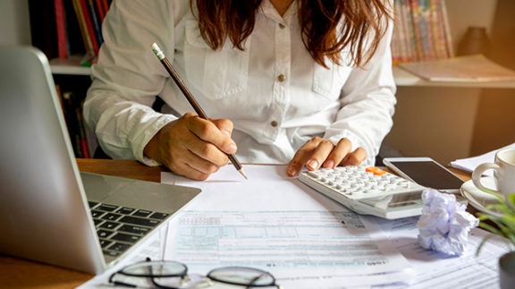 woman writing with a pencil on a document surrounded with a laptop, calculator and glasses