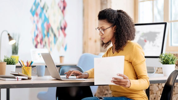 woman sitting at a table working on a laptop and holding papers