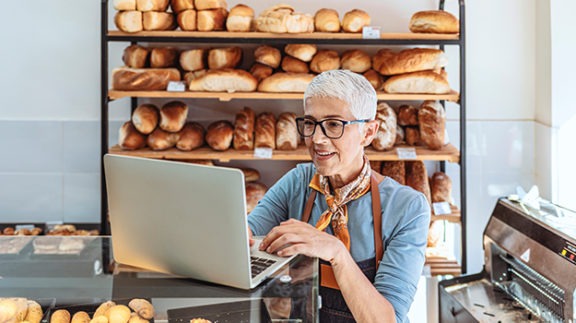 short haired woman on laptop in a bakery surrounded by bread