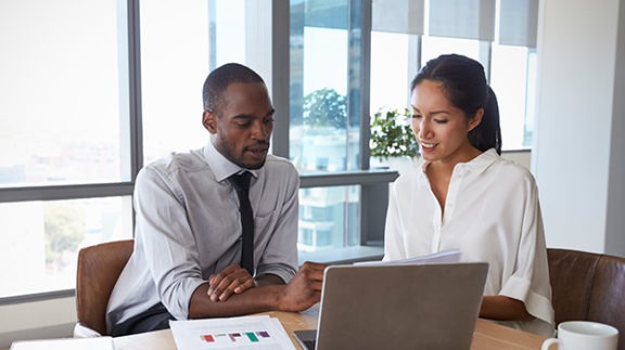 two professionals sitting at a table reviewing documents in front of a laptop